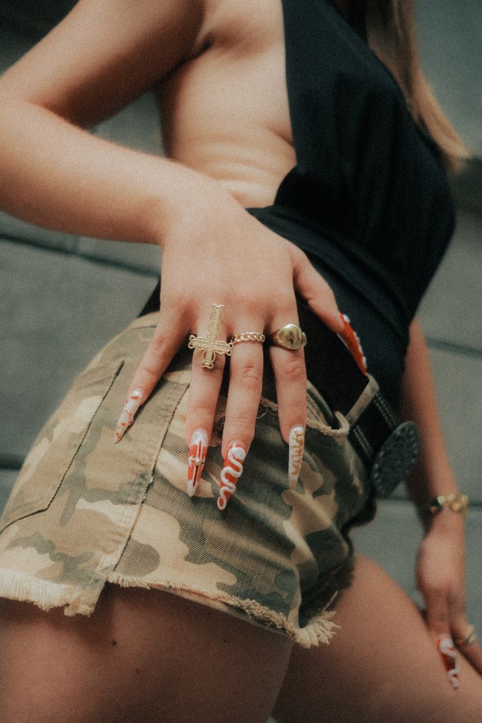 services-02 Close-up of a woman's hand with long acrylic nails and rings, wearing camouflage shorts.